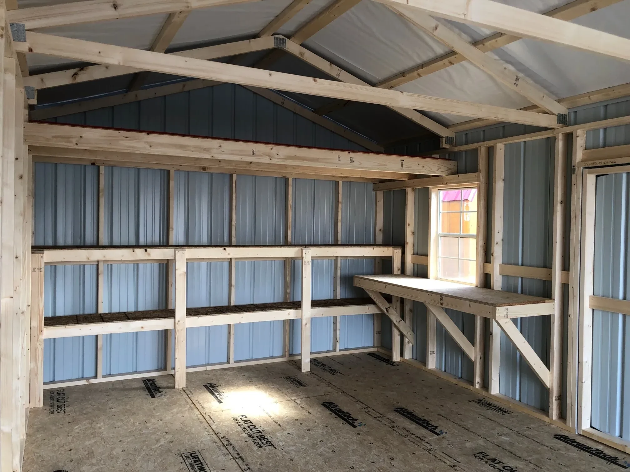 The inside of a metal garage showing a workbench, double shelves, a loft, and a window