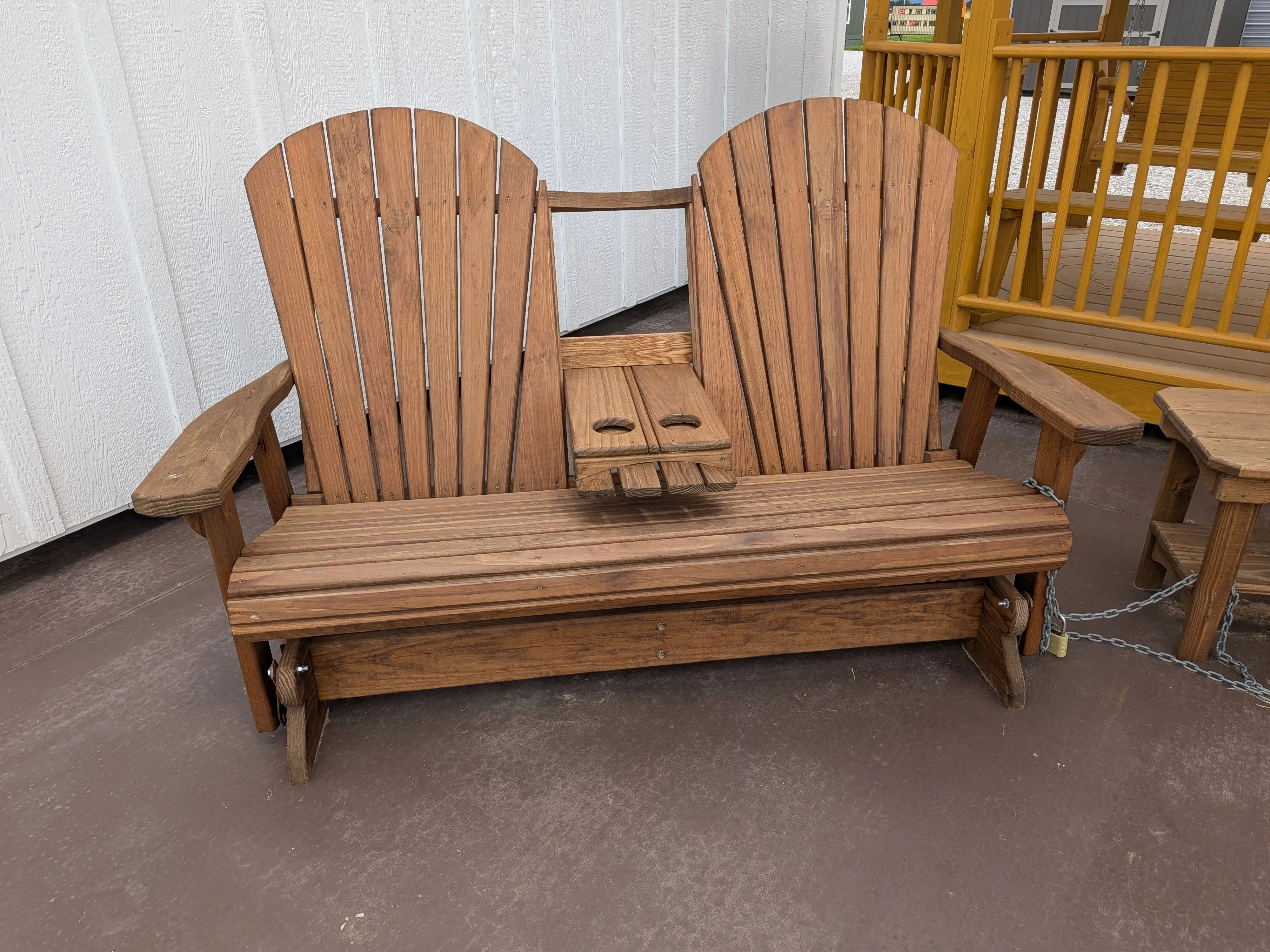a bench that glides back and forth sitting on a concrete pad. It is wood and is stained and sealed. It has a center console