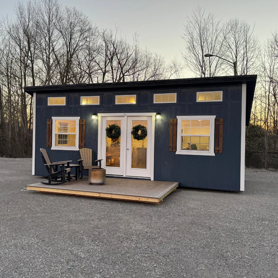 A dark blue studio shed with glass patio doors and many windows and a porch with outdoor furniture