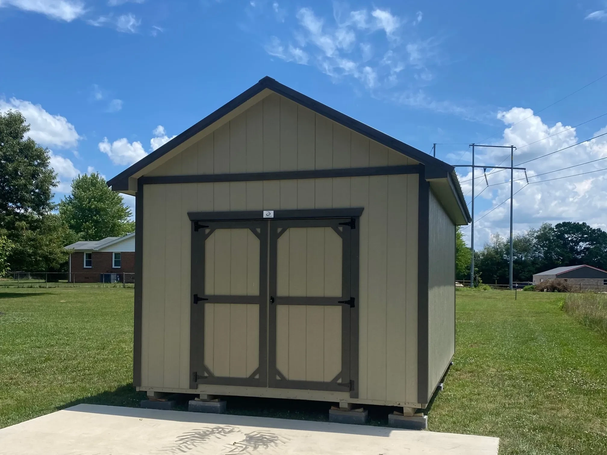 a tan and brown shed with double doors in the end sitting in a yard on a sunny day