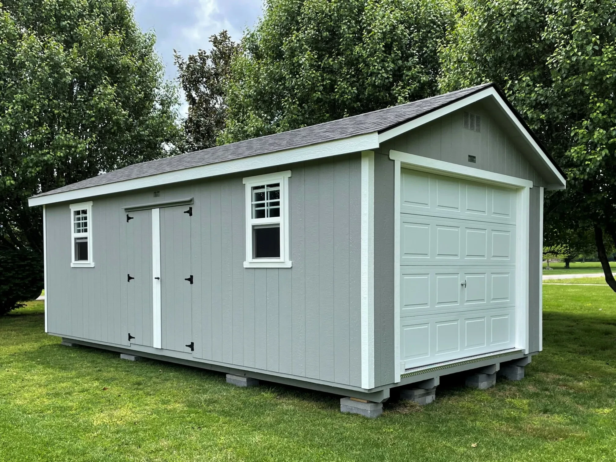 a garage shed with a garage door in the end and double doors and two windows in the side with gray siding and a shingle roof