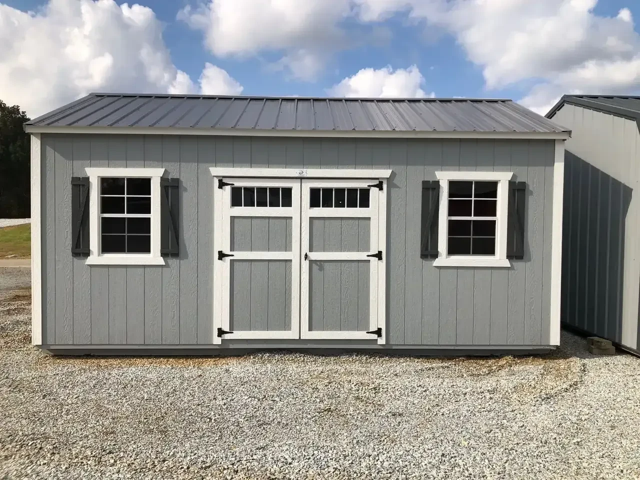 a gray garden shed that has two windows with shutters and double doors with transom windows