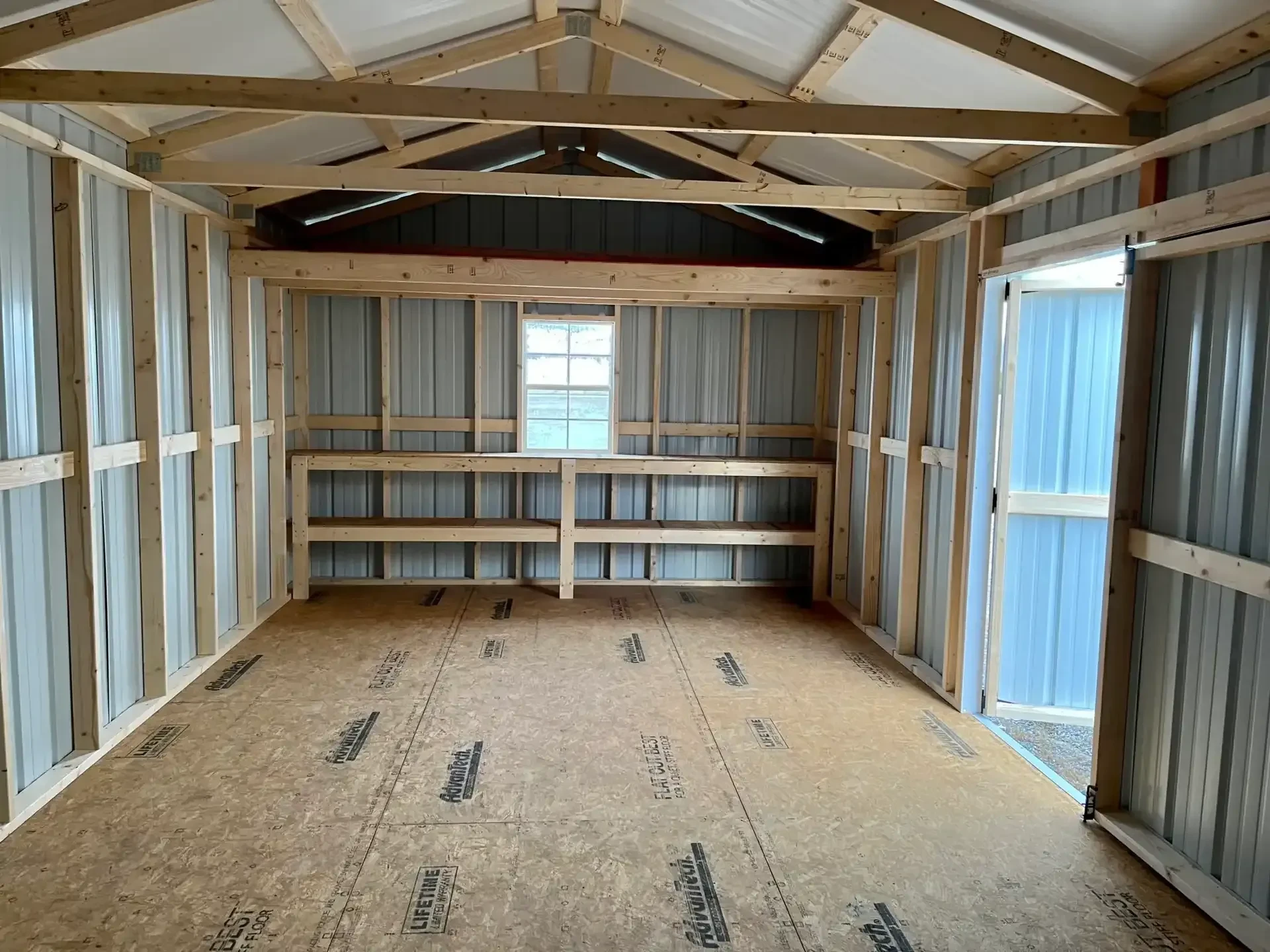 inside a metal garage showing a 4' loft, a window, and double shelves along the back wall, with one of the double doors open