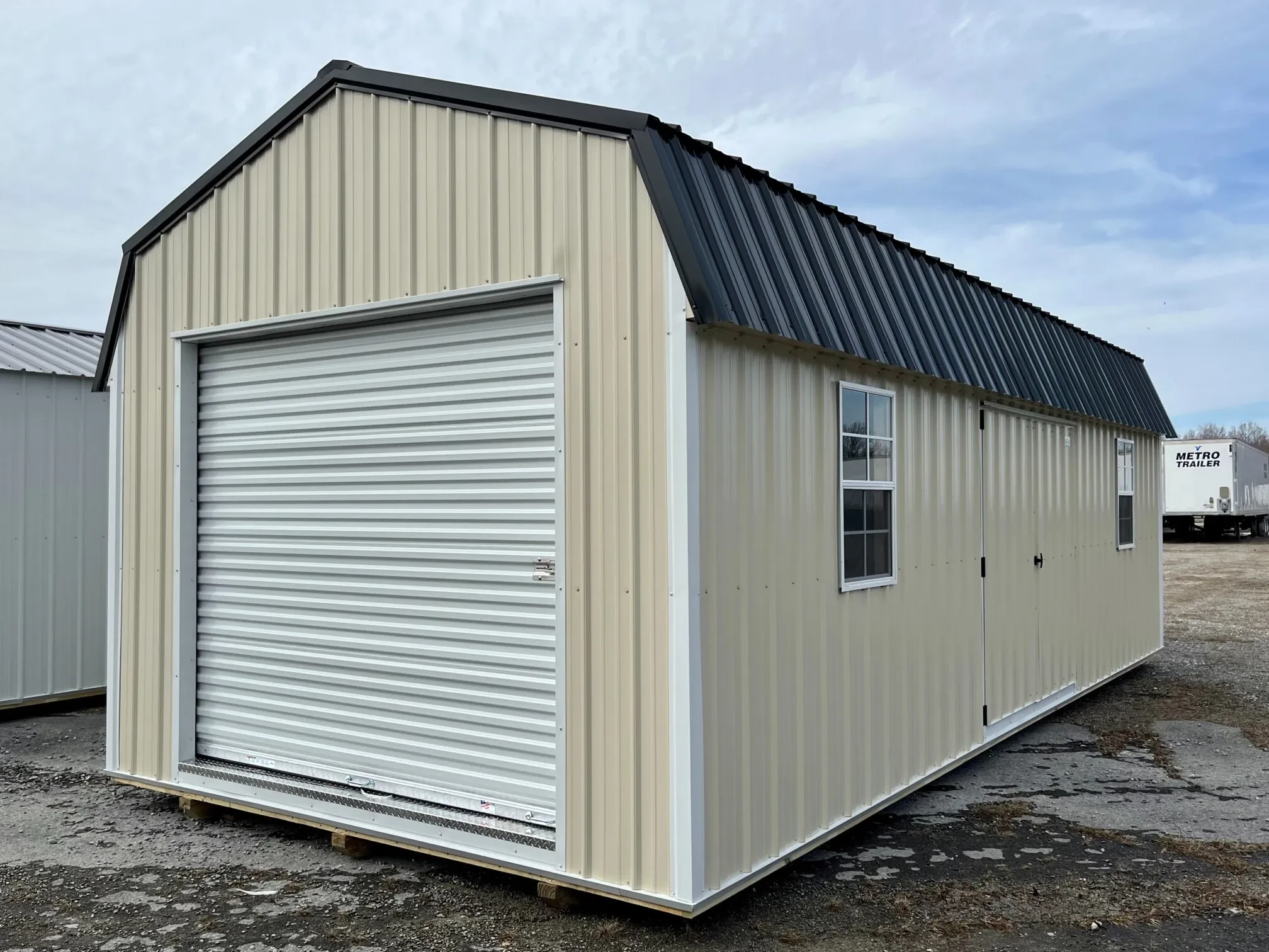 a long stone colored lofted shed with a garage door in the end and double doors and two windows in the side