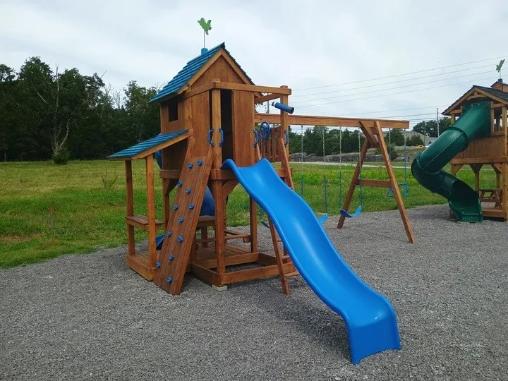 A dark stained wood playhouse playset with blue roof, swings, slide, and rock wall fixtures.