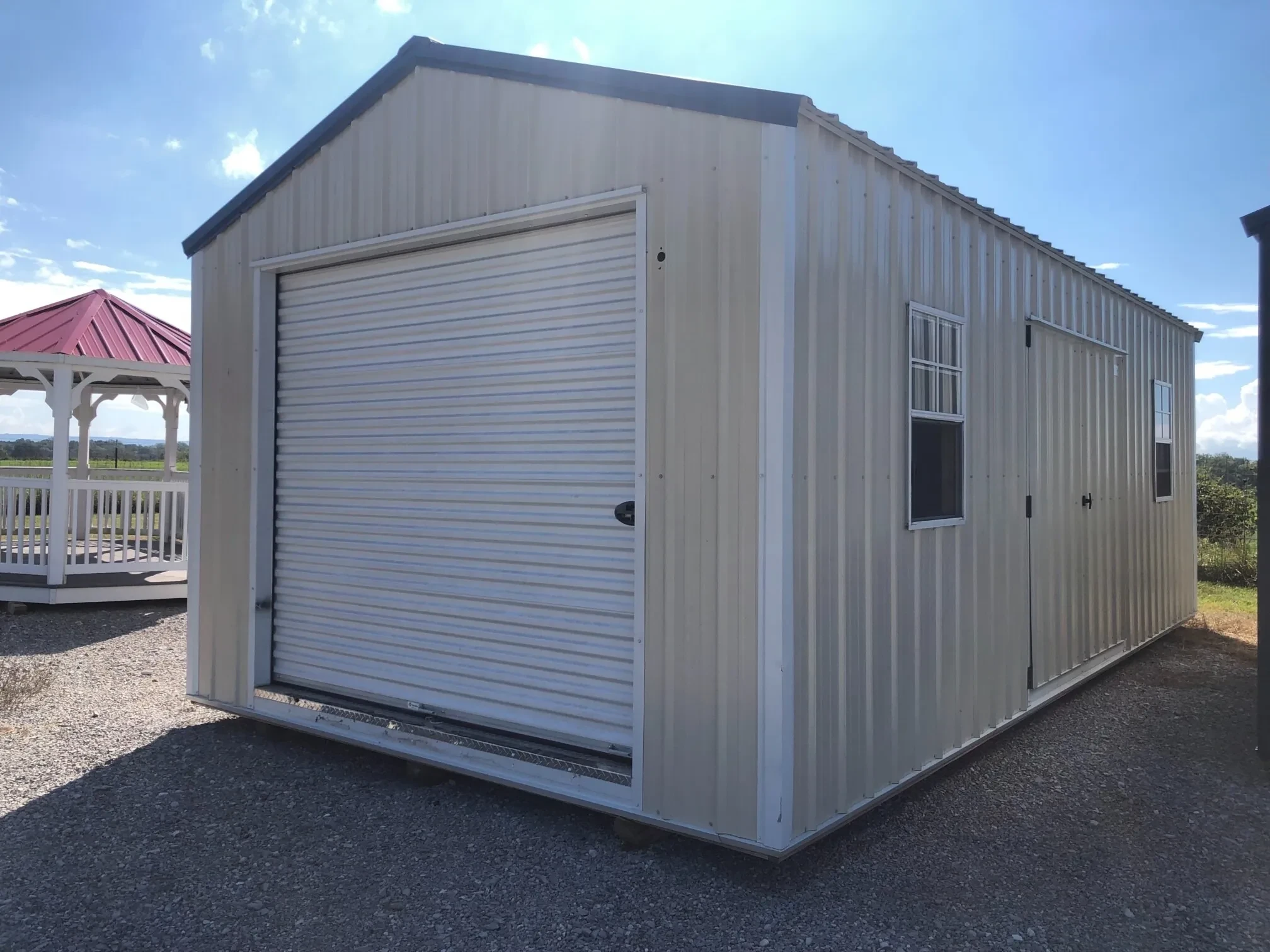 A stone colored metal shed with a garage door on the end, two windows and double doors in the side.