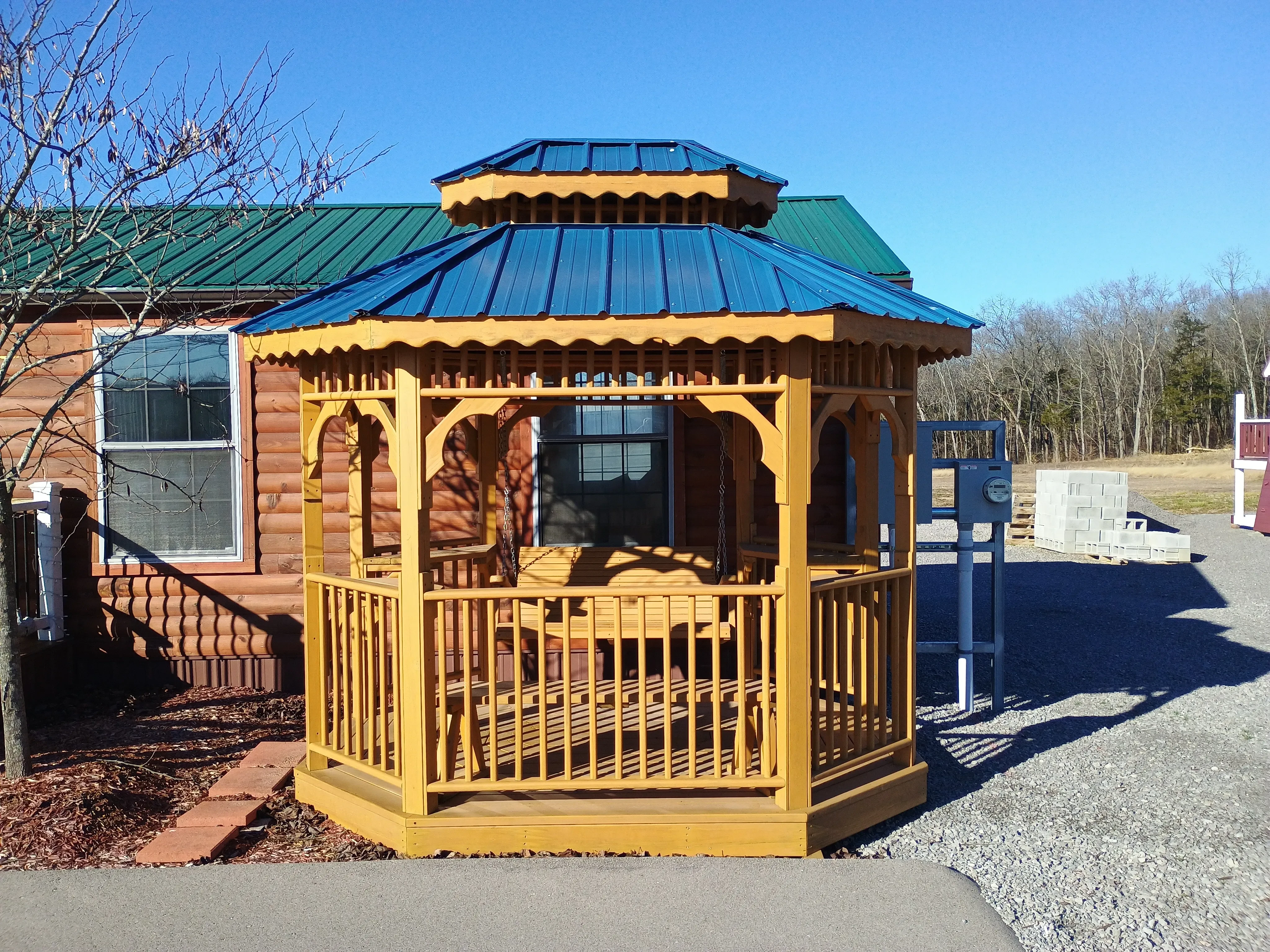 a honey gold stained wood teahouse with posts and railings, it has a bench inside and a blue metal roof