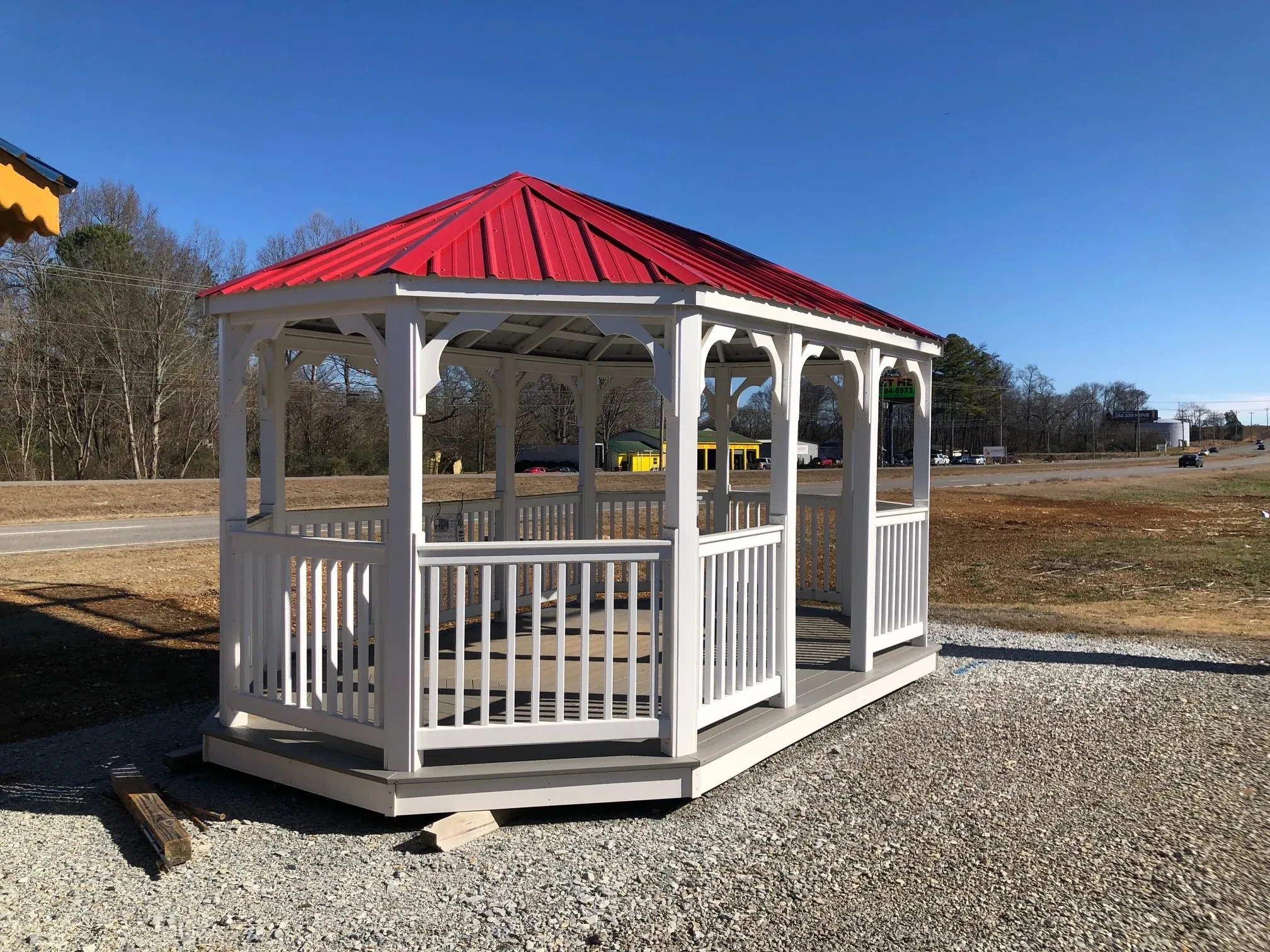 the front of a large white vinyl gazebo with posts, railings, and decorative trim. It has a red metal roof
